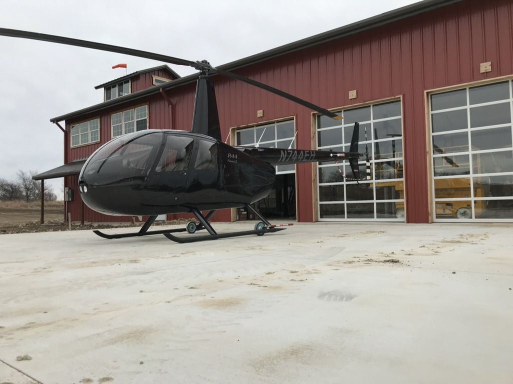 Black R44 helicopter parked in front of The Helo Station's red hangar and glass bay doors